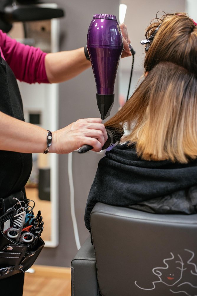 Hairdresser blow-drying and brushing a client’s hair in a salon using professional styling tools.